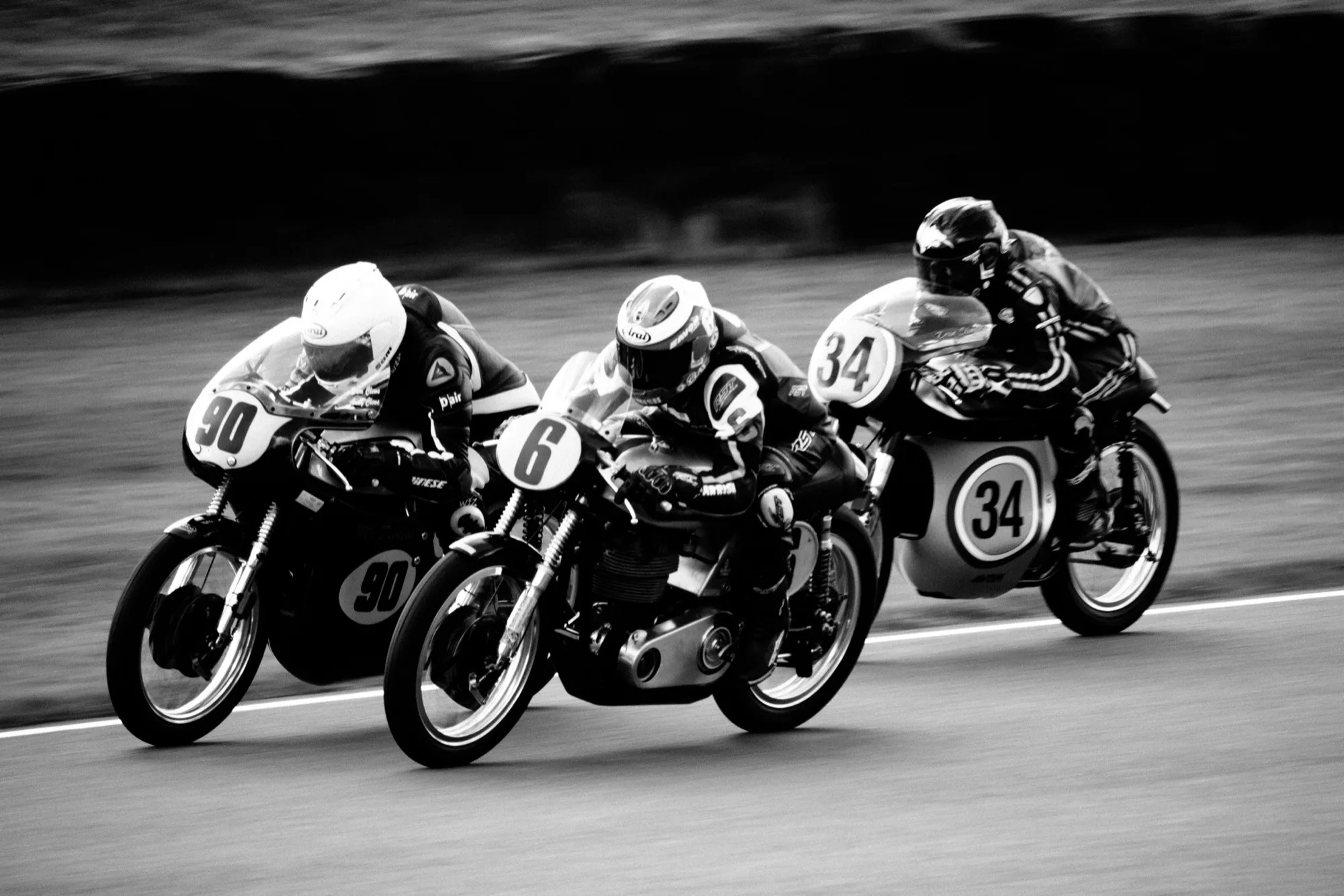 Three vintage racing motorcycles at full lean into a corner, past the braking point, the line is already committed